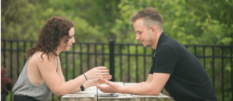 Man and Woman sitting at a table outside in a park like area. Man is reaching for woman's hands, woman is beginning to place her hand in his.