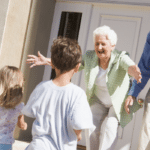 Two children, one boy one girl are running toward two elderly people, one woman, one man while standing on the front porch of a house. The woman is crouch down with her hands out towards the little girl running towards her. Both the elderly woman and the elderly man have smiles on their faces.