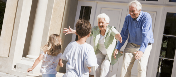 Two children, one boy one girl are running toward two elderly people, one woman, one man while standing on the front porch of a house. The woman is crouch down with her hands out towards the little girl running towards her. Both the elderly woman and the elderly man have smiles on their faces.